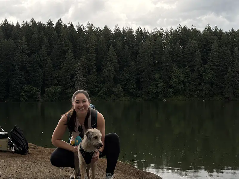 Dr. Hannah Young crouching beside her scruffy tan dog on a lakeside trail, with tall evergreen trees behind them